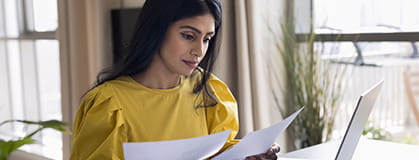 A woman doing accounting paperwork at home, sitting at table with calculator and laptop, reviewing bills