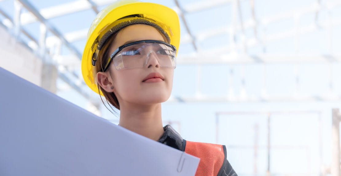Engineer on a construction site holding a blueprint while wearing a safety helmet and goggles.